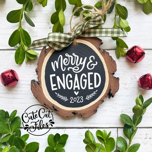 May include: A wooden Christmas ornament with a black chalkboard surface that says "Merry & Engaged 2023". The ornament is decorated with a green and white plaid ribbon and a brown twine hanger.