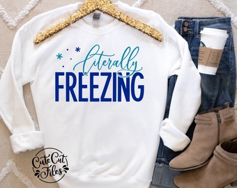 May include: White sweatshirt with blue and teal text that reads "* * literally FREEZING". The sweatshirt is on a hanger and is styled with jeans and brown boots.