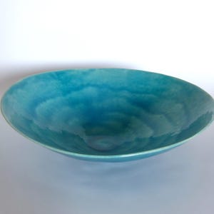 Op de afbeelding: Een turquoise keramische kom met een glanzende afwerking. De kom heeft een breed, ondiep ontwerp en een gladde, gebogen rand. De binnenkant van de kom heeft een gemarmerd effect, met lichtere en donkerdere tinten turquoise.