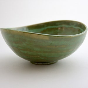 Può includere: Una ciotola in ceramica verde con un bordo ondulato e una sottile lucentezza metallica. La ciotola ha una forma unica e organica, perfetta per servire spuntini o dessert.