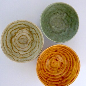 May include: Three ceramic bowls in shades of green, yellow, and tan. Each bowl has a spiral design in the center. The bowls are arranged on a white surface. The bowls are handmade and have a rustic appearance.