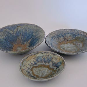 Może przedstawiać: Trzy ceramiczne miski z niebiesko-beżową glazurą. Miski różnią się rozmiarem, największa jest owalna, a najmniejsza okrągła. Glazura ma teksturowany, organiczny wygląd, z niebieskimi i beżowymi smugami.