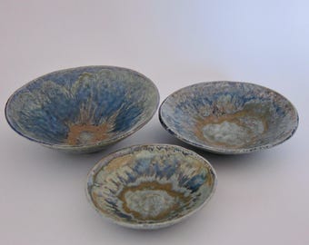 May include: Three ceramic bowls with a blue and tan glaze. The bowls vary in size, with the largest being oval-shaped and the smallest round. The glaze has a textured, organic appearance, with streaks of blue and tan.