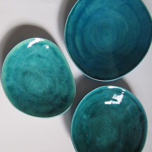 May include: Three turquoise ceramic plates with a glossy finish. The plates are different shapes, including a rounded oval and two circular plates. The plates have a textured surface and are arranged on a white background.