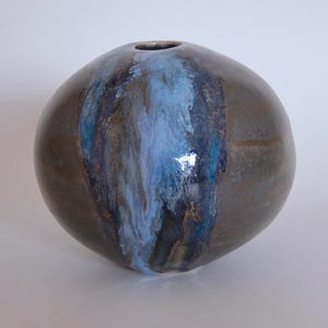 Puede incluir: Un jarrón de cerámica redondo con una base marrón oscuro y una franja vertical de esmalte azul y blanco. El jarrón tiene una pequeña abertura en la parte superior y un acabado liso y brillante. La forma general es esférica.