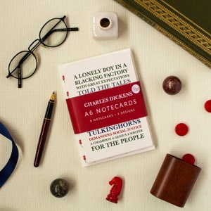 May include: A set of 8 A6 notecards featuring a quote from Charles Dickens, "A lonely boy in a blacking factory with great expectations told the tales." The quote is printed in red and black on a white background. The notecards are packaged in a red, white, and black box with the title "Charles Dickens A6 Notecards" printed on the front.
