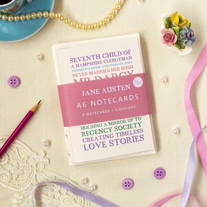 May include: A set of 8 Jane Austen A6 notecards featuring the text "Seventh child of a Hampshire clergyman, supportive sister, affectionate aunt, never married her Irish Mr. Darcy." The notecards are packaged in a pink and white box with the text "Holding a mirror up to Regency society, creating timeless love stories."