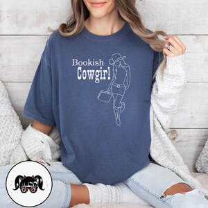 May include: A navy blue t-shirt with a white outline of a woman wearing a cowboy hat and holding a suitcase. The text "Bookish Cowgirl!" is printed on the shirt.