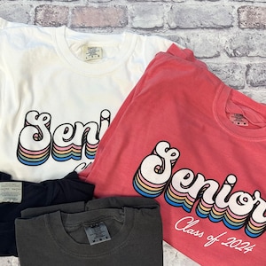 Puede incluir: Tres camisetas con la palabra "Senior" en una fuente retro arco&iacute;ris. La camiseta rosa dice "Class of 2024" debajo de la palabra "Senior".