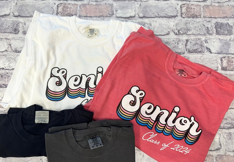 Puede incluir: Tres camisetas con la palabra "Senior" en una fuente retro arco&iacute;ris. La camiseta rosa dice "Class of 2024" debajo de la palabra "Senior".