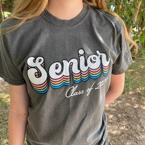 Puede incluir: Una camiseta gris oscuro con un gr&aacute;fico retro de arco&iacute;ris que dice "Senior" y "Class of 2023" en texto blanco.