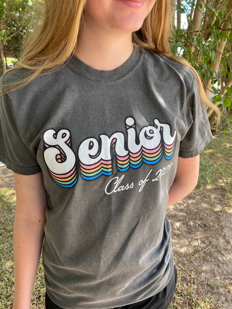 Puede incluir: Una camiseta gris oscuro con un gr&aacute;fico retro de arco&iacute;ris que dice "Senior" y "Class of 2023" en texto blanco.