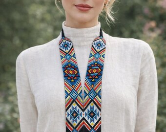 May include: A colourful beaded necklace with an eagle motif. The necklace has a black base with a detailed eagle head and geometric patterns in blue, red, yellow, and white. The necklace is worn over a white turtleneck.