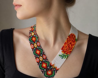 May include: A beaded necklace with a floral design. The necklace features a white band with an orange flower and a patterned band with red, green, yellow, and orange geometric shapes. The necklace is worn on a person wearing a black top.
