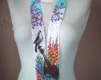 May include: A long, beaded necklace with a blue and white background. The necklace features a beaded design of birds and flowers. The necklace is on a white mannequin.