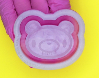 May include: A pink silicone mold shaped like a bear with a dripping face. The mold is held in a gloved hand against a yellow background.