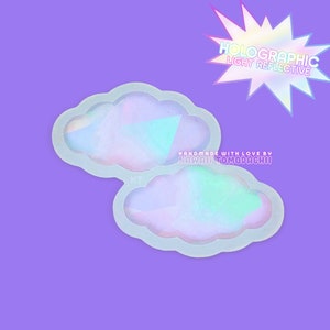 Op de afbeelding: Twee regenboogkleurige, holografische siliconen mallen in de vorm van wolken. De mallen zijn lichtreflecterend en hebben een regenboogeffect. De tekst "HOLOGRAPHIC LIGHT REFLECTIVE" staat in een stervorm boven de mallen. De tekst "HANDMADE WITH LOVE BY KAWAII TOMODACHI" staat onder de mallen.