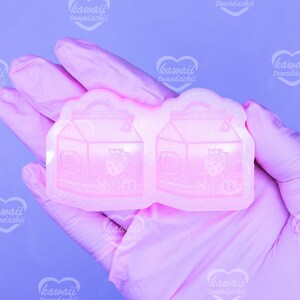 May include: A light pink silicone mold featuring two strawberry milk carton designs. Each carton has a strawberry graphic and the word "milk" in Japanese. The mold is held by a person wearing pink gloves.