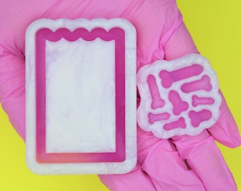 May include: Two pink silicone molds, one shaped like a rectangle with a wavy edge and the other shaped like a cluster of small bones.