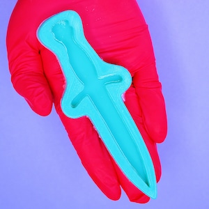 Könnte beinhalten: Eine türkisblaue Silikonform in Form eines Dolches. Die Form wird in einem rosa Latexhandschuh gehalten.