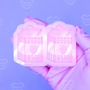 May include: A pink resin mold in the shape of a strawberry milk carton. The carton design includes a strawberry graphic, text, and a straw. The mold is held by a gloved hand against a purple background.