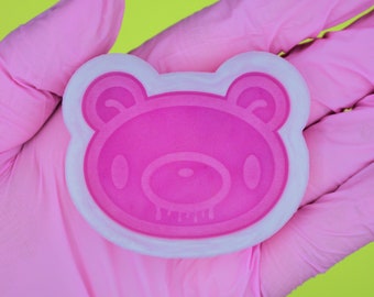 May include: A pink silicone mold shaped like a bear with a white outline. The bear has a simple design with two eyes and a dripping mouth.
