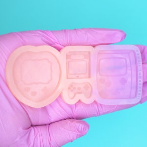 May include: A pink silicone mold with three sections. The sections are shaped like a heart with a frame inside, a handheld video game console, and a handheld game console with a screen and buttons. The mold is for making resin crafts.