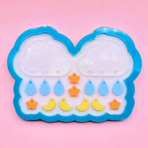 May include: A blue cloud-shaped resin mold with a clear center, surrounded by blue trim. The mold contains blue teardrops, orange stars, and yellow crescent moon shapes. The mold is designed for crafting.