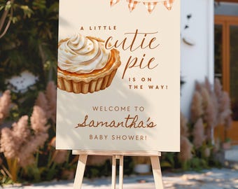 May include: A rectangular sign on a wooden easel featuring a watercolor illustration of a small pie with whipped cream. The text reads A LITTLE cutie pie IS ON THE WAY! WELCOME TO Samantha's BABY SHOWER! with orange gingham bunting at the top.