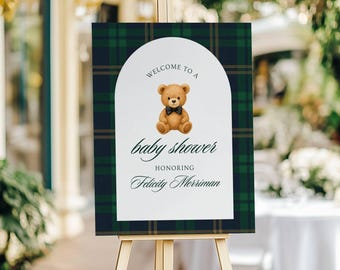 May include: A rectangular sign with a dark green and navy blue plaid border featuring a white arched center. The text reads Welcome to a baby shower honoring Felicity Merriman, accompanied by an illustration of a brown teddy bear with a black bow tie.