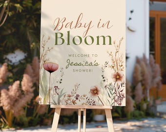 May include: A rectangular welcome sign on a wooden easel featuring the text Baby in Bloom and Welcome to Jessica's Shower. The cream-colored sign is decorated with watercolor illustrations of wildflowers in shades of pink, brown, and green.