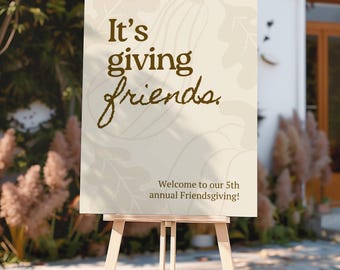 May include: A rectangular cream-colored sign on a wooden easel featuring the dark brown text It's giving friends. and Welcome to our 5th annual Friendsgiving! The background displays faint line drawings of pumpkins and oak leaves in a light tan color.