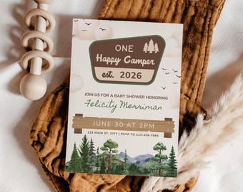 May include: A rectangular baby shower invitation card featuring a brown badge with the text ONE Happy Camper est. 2026, followed by the name Felicity Merriman, the date June 30 at 2PM, and address details, set against a forest and mountain landscape.
