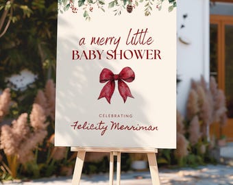 May include: A rectangular sign on a wooden easel featuring a cream background, a watercolour holly and pinecone border, and a dark red bow. The text reads: a merry little BABY SHOWER CELEBRATING Felicity Merriman in dark red script and block lettering.