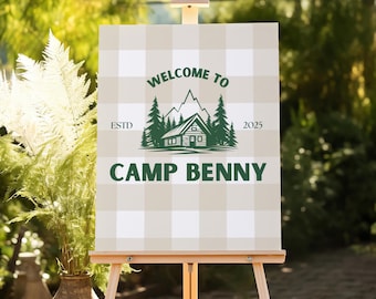 Op de afbeelding: Een rechthoekig bord op een houten ezel met een beige en wit ruitpatroon. Het bord toont een donkergroene afbeelding van een hut, dennenbomen en bergen, met de tekst "WELCOME TO", "ESTD 2025" en "CAMP BENNY".