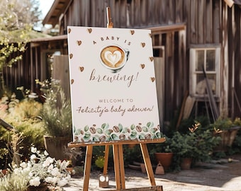 May include: A rectangular white sign on a wooden easel featuring a coffee cup illustration with heart-shaped latte art. The text reads A BABY IS brewing! WELCOME TO Felicity's baby shower! The bottom border displays coffee beans and green leaves.