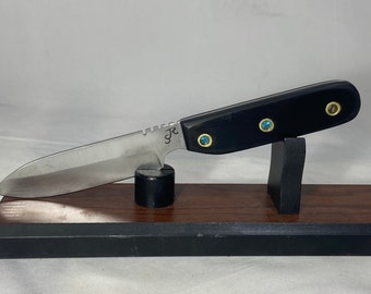 Könnte beinhalten: Ein Messer mit schwarzem Griff, silberner Klinge und blauen Edelsteinen am Griff. Das Messer befindet sich auf einem Holzständer.