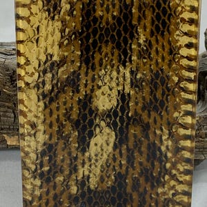 May include: A rectangular object featuring a detailed, repeating snakeskin pattern in shades of dark brown, black, and tan. The surface displays a textured, scale-like appearance with a glossy finish, showcasing a naturalistic reptile skin motif.