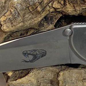 May include: A folding pocket knife with a metallic silver blade featuring a laser-engraved graphic of a snake head with an open mouth and a protruding tongue. The knife handle is partially visible, showing a metallic finish and a screw fastener.