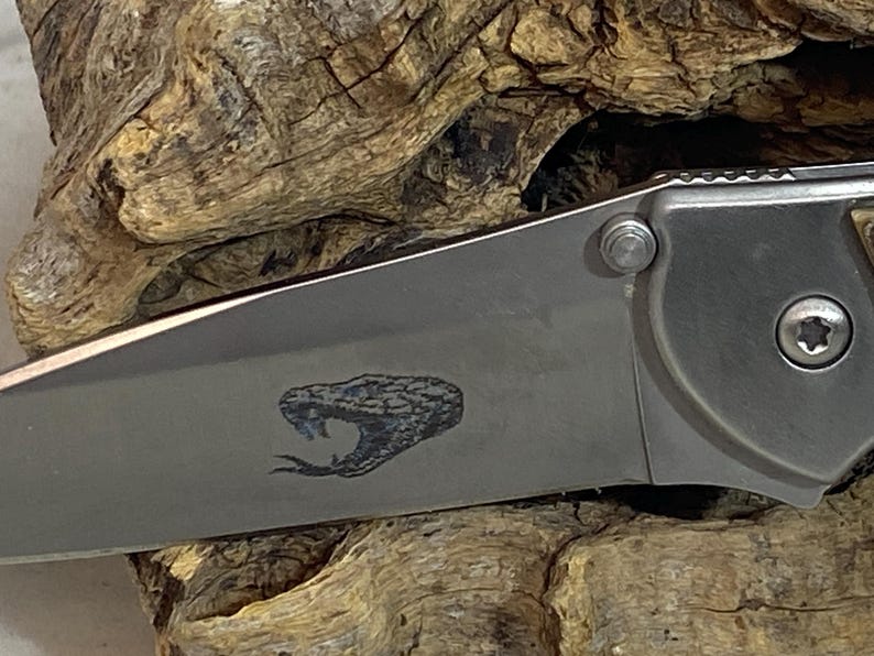 May include: A folding pocket knife with a metallic silver blade featuring a laser-engraved graphic of a snake head with an open mouth and a protruding tongue. The knife handle is partially visible, showing a metallic finish and a screw fastener.
