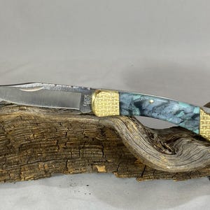 以下が含まれることがあります： Buck 110の刻印が入った銀色の金属製ブレードを持つ折りたたみ式ポケットナイフ。ハンドルは両端に質感のある金色の金属製ボルスターがあり、中央には青、黒、白のマーブル模様のアクリル製グリップセクションが施されています。