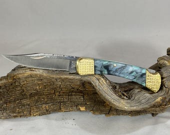 Könnte beinhalten: Ein Klappmesser mit einer silberfarbenen Metallklinge, die den Schriftzug Buck 110 trägt. Der Griff hat an beiden Enden strukturierte, goldfarbene Metallbacken und in der Mitte ein Griffstück aus marmoriertem blauem, schwarzem und weißem Acryl.