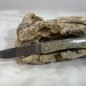 May include: A folding pocket knife with a polished, light green handle and a silver blade. The knife is open, revealing the sharp edge. The handle is attached with a silver rivet. The knife rests on a piece of weathered wood.