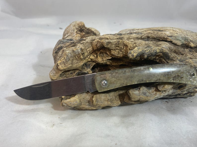 May include: A folding pocket knife with a polished, light green handle and a silver blade. The knife is open, revealing the sharp edge. The handle is attached with a silver rivet. The knife rests on a piece of weathered wood.