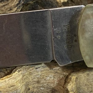 May include: Close-up of a pocket knife blade with the text "CASE XX" etched into the metal. The blade is silver and the handle is a light tan color. The knife is resting on a textured wooden surface.