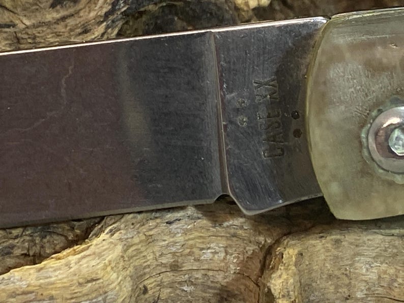 May include: Close-up of a pocket knife blade with the text "CASE XX" etched into the metal. The blade is silver and the handle is a light tan color. The knife is resting on a textured wooden surface.