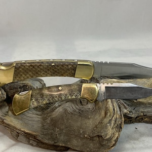 May include: Two folding pocket knives with metallic blades, brass bolsters, and textured brown handles featuring a scale-like pattern. The knives are positioned one above the other, showcasing a classic lock-back design with visible brass pins.