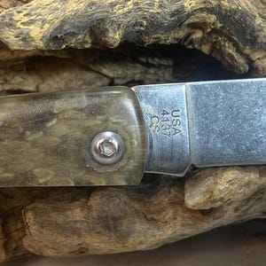 May include: Close-up of a folding knife with a silver blade and a handle with a clear, textured appearance. The blade is marked "USA 4137 C.S.". The knife is set against a wooden background.