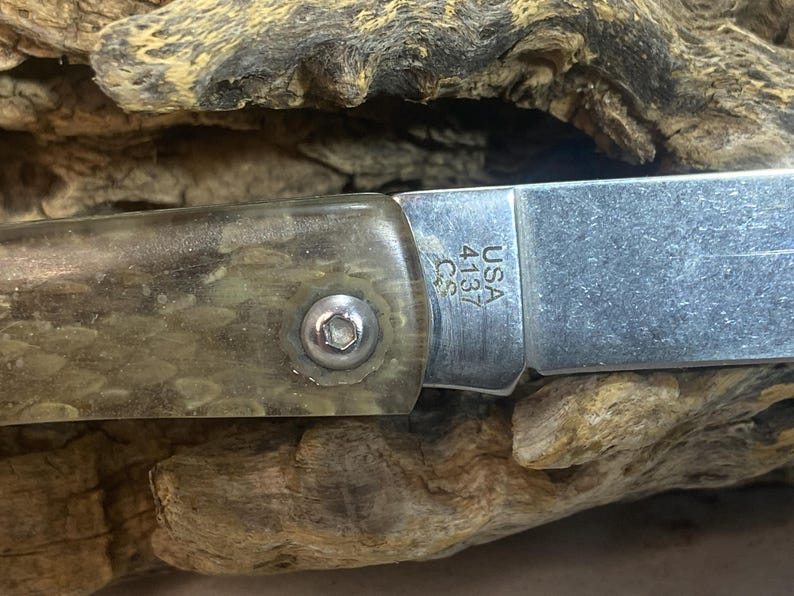 May include: Close-up of a folding knife with a silver blade and a handle with a clear, textured appearance. The blade is marked "USA 4137 C.S.". The knife is set against a wooden background.