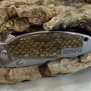 May include: A closed folding pocket knife with a metallic silver handle and a translucent amber-colored honeycomb patterned inlay. The handle features the text Kershaw SpeedSafe USA and a small circular hole at the end for a lanyard attachment.